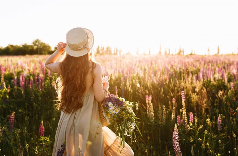 Young woman in straw hat and dress with bouquet of lupine flowers, back view in sunset field.